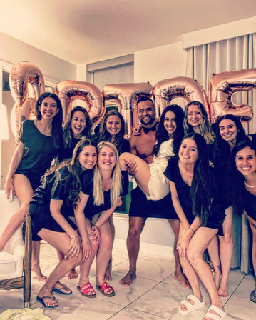 Bachelorette party: smiling women and a man pose indoors as the bride-to-be in white is lifted in front of large rose-gold "BRIDE" balloons.