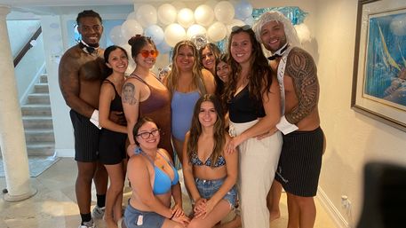 Group of friends in swimwear and summer outfits smiling at camera indoors in front of white and light-blue balloon backdrop, two men in bow ties and striped shorts flanking the group at a casual pool-themed celebration.