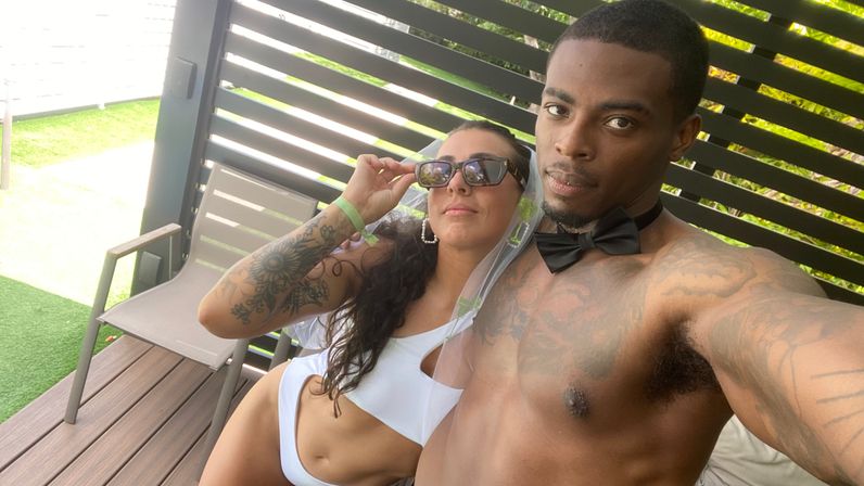 Couple selfie in an outdoor cabana: shirtless man wearing a black bow tie collar and a woman in a white bikini and sunglasses with a floral arm tattoo, wooden slatted privacy screen and grassy yard behind them.