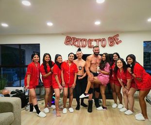 Bachelorette party at home: bride-to-be in a pink robe poses with friends in matching red pajama sets and two shirtless male dancers under a "Bride to Be" balloon banner in a living room.