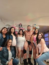 Group photo at an indoor home celebration: six women posing with a shirtless male entertainer in dance briefs, silver fringe backdrop, cowgirl boot and star balloons, disco ball and festive party vibe.