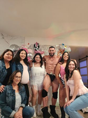 Shirtless male entertainer in black briefs and knee pads posing with a group of smiling women at an indoor living-room party with silver fringe backdrop, pink and silver balloons and novelty cowboy-boot balloon.