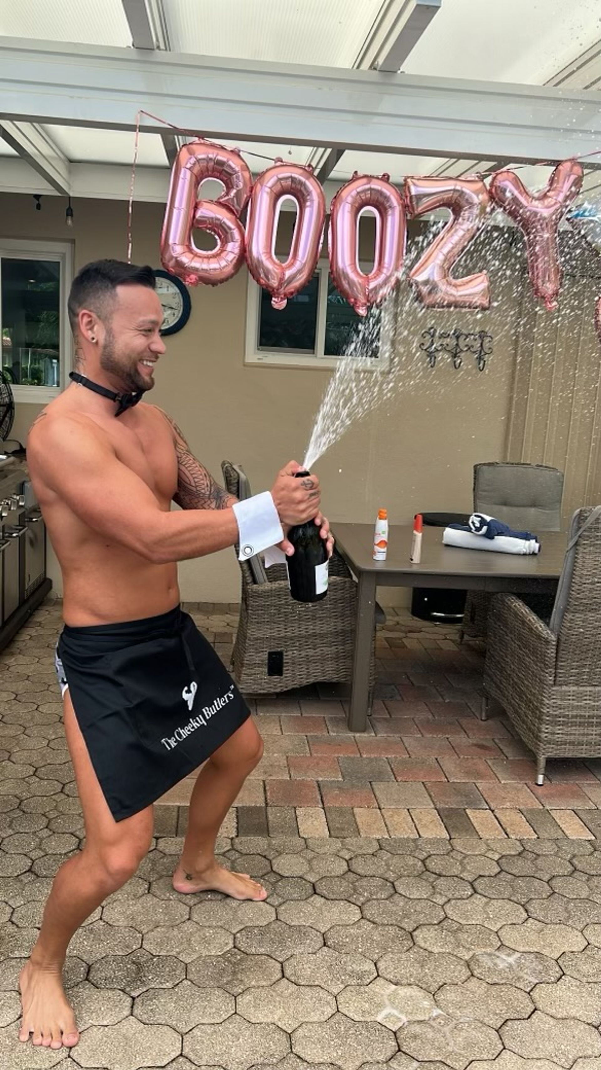 Shirtless man in a black half-apron popping a champagne bottle, bubbly spraying under rose-gold letter balloons on a covered backyard patio with wicker furniture — festive poolside party vibe.