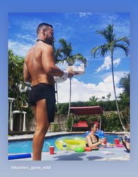 Shirtless server in black shorts carrying plates at a sunny tropical backyard pool with palm trees, colorful inflatables, drinks on the deck and a woman lounging in the water under a bright blue sky.