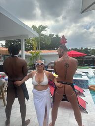 Playful poolside group: woman in white bikini and heart-shaped sunglasses smiles between two men in aprons and thong swimwear at a tropical backyard pool with palm trees and red umbrellas.