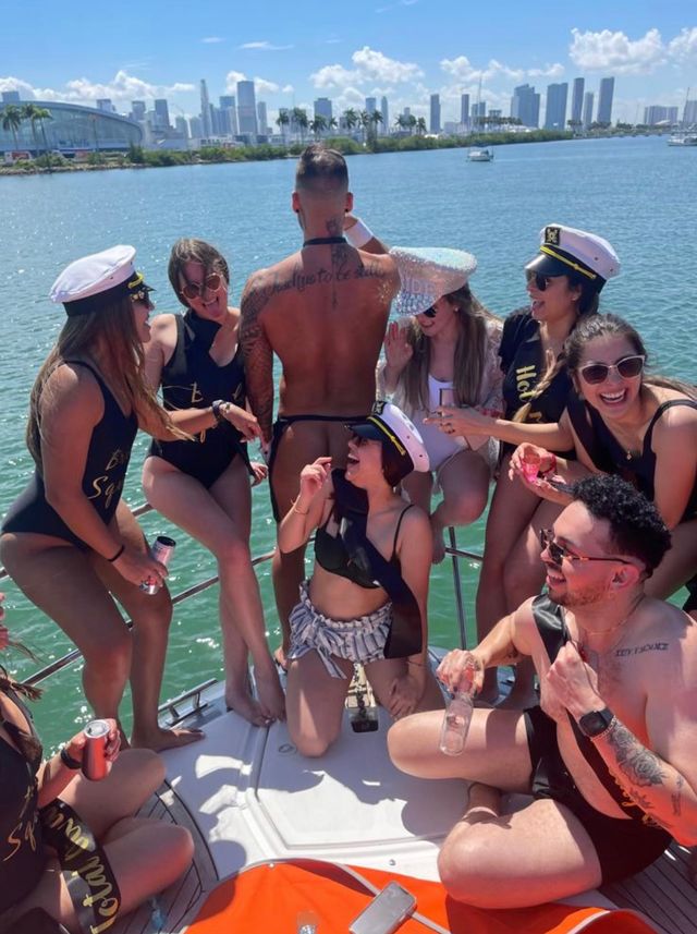 Yacht bachelorette party on a sunny day by a waterfront city skyline — friends in captain hats and matching black swimsuits with sashes, laughing and holding drinks around a shirtless man in a thong.