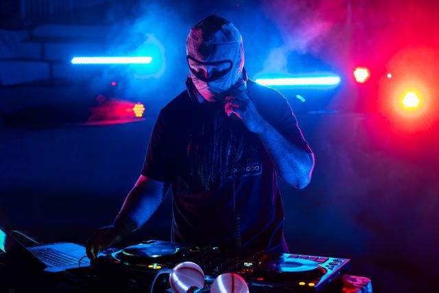 Masked DJ performing a live electronic set, mixing on a controller and laptop amid blue and pink neon lights and stage smoke in a nightclub