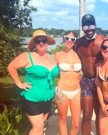 Lakeside summer scene: four people in colorful swimsuits and sunglasses posing under a palm tree by a lake — woman in a green tankini and wide sun hat, woman in a white bikini with heart-shaped sunglasses and a thigh tattoo, a man in patterned swim trunks and cap, and another woman partially visible on the right.