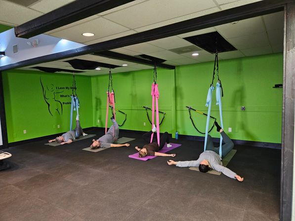 Aerial yoga class in a bright green studio with pink and blue silk hammocks suspended from ceiling beams as participants lie on mats with legs supported in the slings