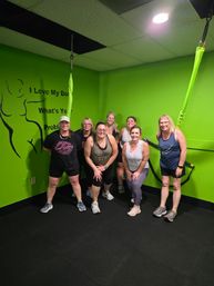 Smiling group of seven women posing in a lime-green fitness studio with suspended green exercise straps, black rubber flooring and motivational wall art.