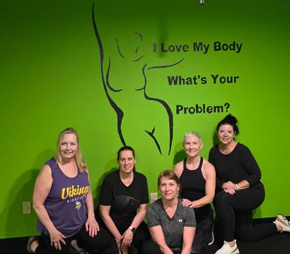 Five women in workout clothes smiling and kneeling in a bright green fitness studio in front of a black outline body mural and text that reads I Love My Body What's Your Problem?