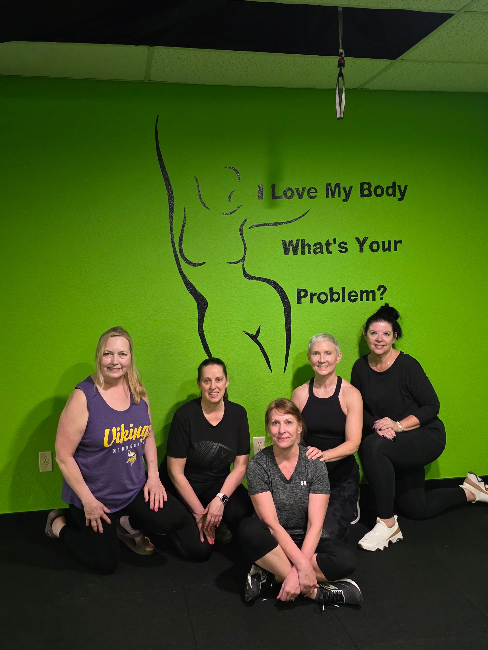 Five women in workout clothes smiling and kneeling in a bright green fitness studio in front of a black outline body mural and text that reads I Love My Body What's Your Problem?