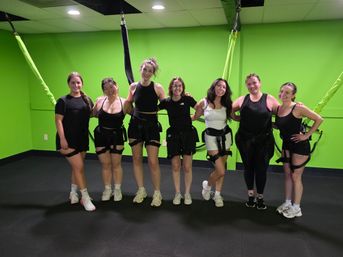Seven women smiling in a bright lime-green indoor fitness studio wearing bungee harnesses attached to the ceiling, posing after an aerial bungee workout class.