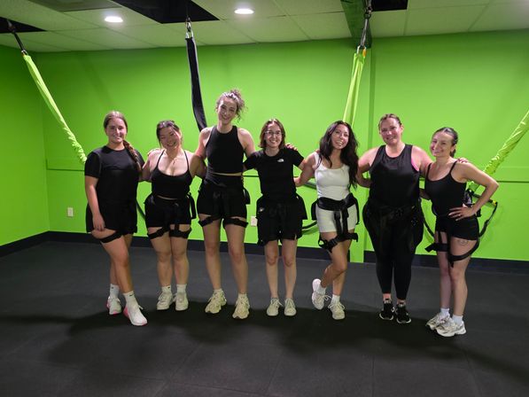 Seven women smiling in a bright lime-green indoor fitness studio wearing bungee harnesses attached to the ceiling, posing after an aerial bungee workout class.