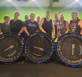 Smiling group of women in a bright green indoor fitness studio standing behind rebounder mini-trampolines, posing after a lively group bounce workout.