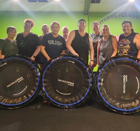 Smiling group of women in a bright green indoor fitness studio standing behind rebounder mini-trampolines, posing after a lively group bounce workout.