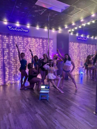 Group of dancers posing around a pole in a purple-lit studio with string lights, mirrored wall, wooden floor and a laptop on a small stool — playful pole-fitness vibe.