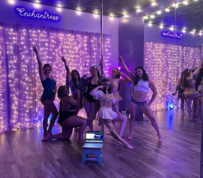 Group of dancers posing around a pole in a purple-lit studio with string lights, mirrored wall, wooden floor and a laptop on a small stool — playful pole-fitness vibe.