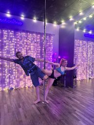 Two people striking a playful pole-dance pose inside a purple-lit pole studio with string lights, neon signage and wood flooring.