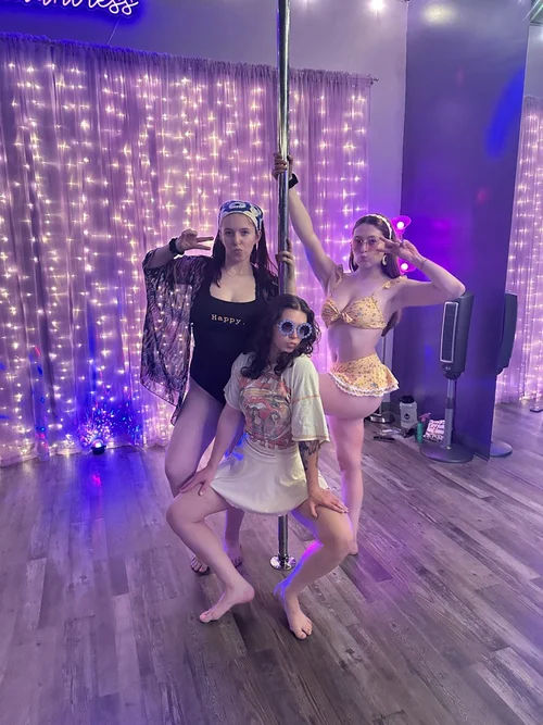 Three dancers posing around a chrome pole in a pole-dance studio with purple fairy lights, wood floor, and colorful outfits, capturing playful fitness and studio vibes.