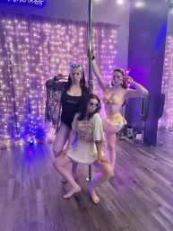 Three dancers posing around a chrome pole in a pole-dance studio with purple fairy lights, wood floor, and colorful outfits, capturing playful fitness and studio vibes.