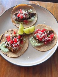 Three grilled steak tacos on warm corn tortillas with green crema, fresh pico de gallo and crumbled cheese, served with lime wedges on a round plate over a wooden table