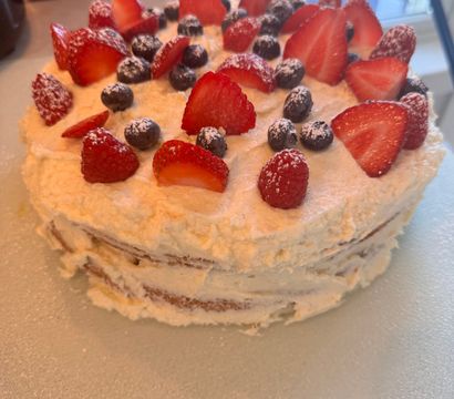 Homemade three-layer vanilla cake with whipped cream frosting topped with sliced strawberries, raspberries and blueberries dusted with powdered sugar on a kitchen countertop.
