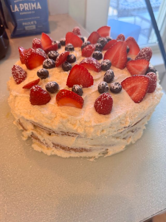 Homemade three-layer vanilla cake with whipped cream frosting topped with sliced strawberries, raspberries and blueberries dusted with powdered sugar on a kitchen countertop.