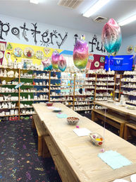 Bright pottery-painting studio with long wooden tables and benches, shelves lined with unpainted ceramic pieces, rainbow metallic balloons floating above, patterned carpet and bowls set at painting stations ready for a kids' party or craft session.