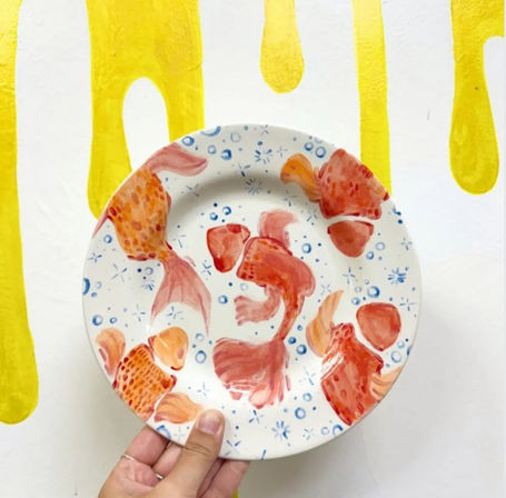 Playful hand-held hand-painted ceramic plate with orange goldfish and blue bubbles on white, photographed against a yellow paint-drip backdrop.