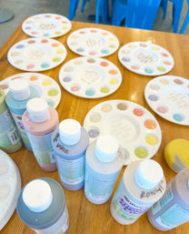 Craft studio table set for a paint party with pastel acrylic bottles, round ceramic palettes dotted with colorful paint samples, a yellow sponge and blue plastic chairs in the background.