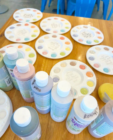 Craft studio table set for a paint party with pastel acrylic bottles, round ceramic palettes dotted with colorful paint samples, a yellow sponge and blue plastic chairs in the background.
