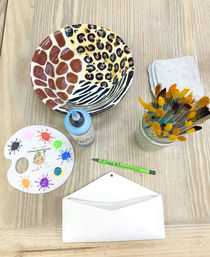 Pottery painting studio table with colorful animal-print ceramic bowl, paint palette with splatters, blue paint bottle, jar of brushes, sponge, and blank ceramic envelope ready to decorate