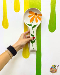 Hand holding a painted ceramic spoon rest with an orange daisy and green stem against a white wall with bold yellow and green paint drips — bright, playful art studio shot