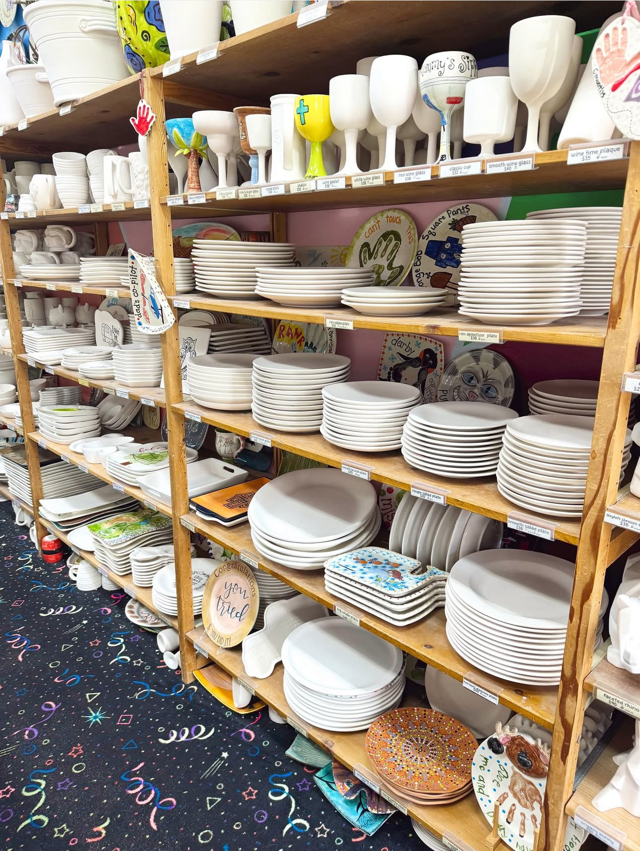 Shelves in a paint-your-own pottery studio packed with stacked blank white ceramic plates, bowls and goblets ready for decorating, with a few colorful finished pieces and patterned carpet below.