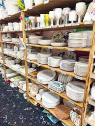 Shelves in a paint-your-own pottery studio packed with stacked blank white ceramic plates, bowls and goblets ready for decorating, with a few colorful finished pieces and patterned carpet below.