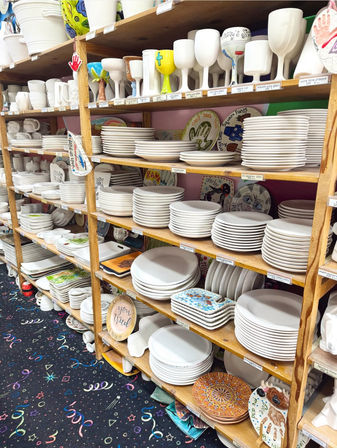 Shelves in a paint-your-own pottery studio packed with stacked blank white ceramic plates, bowls and goblets ready for decorating, with a few colorful finished pieces and patterned carpet below.