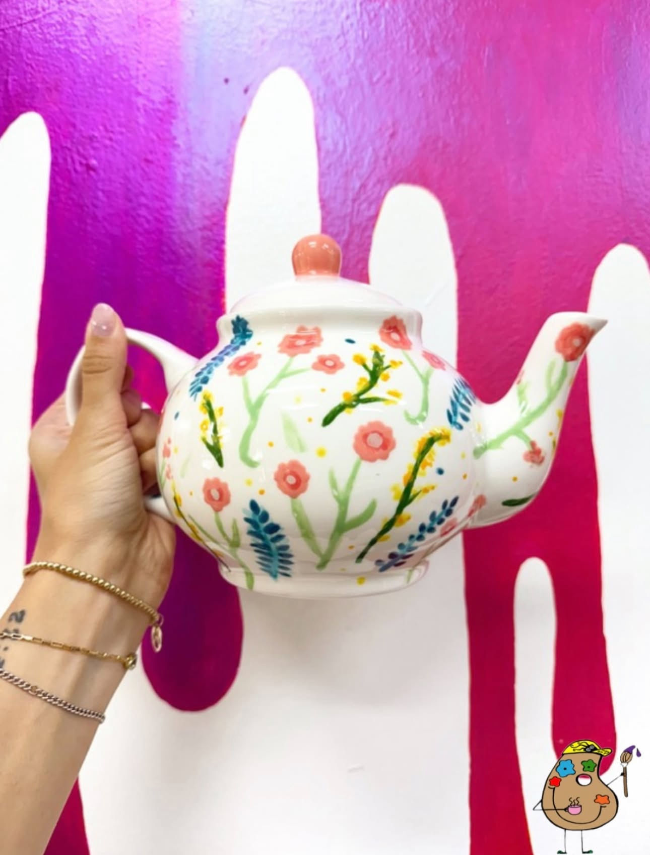 Hand holding a white ceramic teapot hand-painted with pink and yellow flowers and green leaves against a vibrant pink drip mural wall