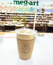 Iced coffee in a plastic cup with cardboard sleeve and straw, sitting on a table inside a colorful paint‑your‑own pottery studio filled with shelves of painted ceramics and decorative plates.