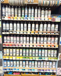 Vibrant rows of labeled squeeze paint bottles with matching color swatches on wooden shelves in a craft supply display, showing pastel, neutral and bright hues for art projects.