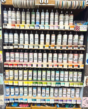 Vibrant rows of labeled squeeze paint bottles with matching color swatches on wooden shelves in a craft supply display, showing pastel, neutral and bright hues for art projects.