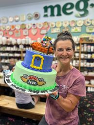 Smiling woman in a pottery painting studio holding a colorful two-tier celebration cake — green and blue tiers with a yellow picture-frame detail, an orange couch and paintbrush topper, and a playful coffee-shop logo for a sitcom-themed party.