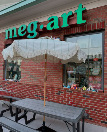 Fringed cream patio umbrella shading a gray picnic table outside a red-brick retail storefront with a bright green sign and a window display of ceramics and colorful figurines.