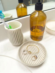 Bathroom vanity scene: striped ceramic toothbrush holder and matching jewelry dish holding rings and a bracelet, amber pump soap bottle and mirror reflection on a white countertop.