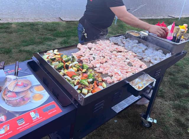 Backyard flat-top griddle cooking diced chicken, shrimp, and colorful mixed vegetables on an outdoor grill station with condiments and a person reaching for squeeze bottles.