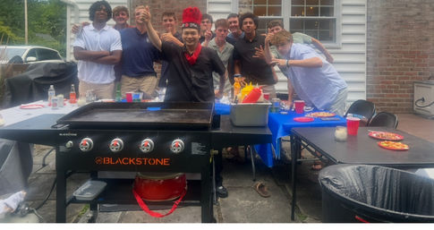Group of friends posing behind a large flat-top griddle on a suburban backyard patio during a casual summer cookout, with condiment bottles, red cups and plates on folding tables.