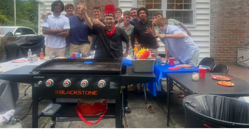 Group of friends posing behind a large flat-top griddle on a suburban backyard patio during a casual summer cookout, with condiment bottles, red cups and plates on folding tables.