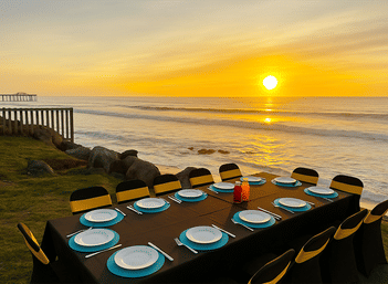 Beachside sunset dining on a grassy bluff: long table with black linen, turquoise chargers and white plates, yellow-banded chairs and condiment bottles overlooking rocky shoreline, waves and a glowing sun with a distant pier.