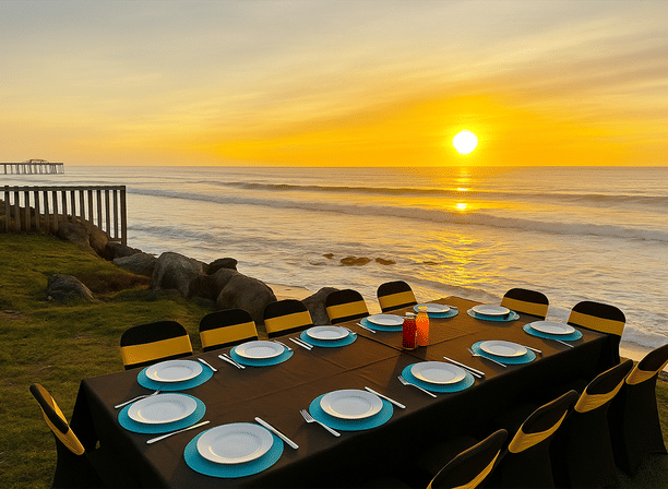 Beachside sunset dining on a grassy bluff: long table with black linen, turquoise chargers and white plates, yellow-banded chairs and condiment bottles overlooking rocky shoreline, waves and a glowing sun with a distant pier.
