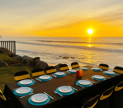 Beachside sunset dining on a grassy bluff: long table with black linen, turquoise chargers and white plates, yellow-banded chairs and condiment bottles overlooking rocky shoreline, waves and a glowing sun with a distant pier.